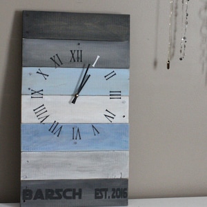 May include: A rectangular wooden wall clock with a rustic design. The clock face features Roman numerals and black clock hands. The clock is made of horizontal wooden planks painted in shades of gray and blue. The text "BARSCH EST. 2016" is printed at the bottom.