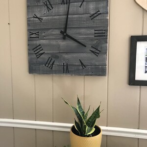 K&ouml;nnte beinhalten: Eine graue Holzuhr mit r&ouml;mischen Ziffern an einer wei&szlig;en Wand. Die Uhr hat schwarze Zeiger und h&auml;ngt &uuml;ber einer Topfpflanze.
