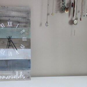 May include: A rustic wooden clock with Roman numerals and a blue, green, and gray color scheme. The clock is mounted on a wall and has the name "Emmanuelidis" written on the bottom.