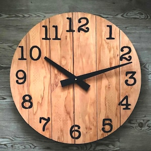 May include: A round wooden clock with black numerals and hands. The clock face is made of light brown wood planks arranged vertically. The numbers are in a bold, sans-serif font. The hands are black and point to 2:10.