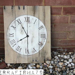May include: Square wooden clock with a white face and black Roman numerals. The clock hands are black. The clock is made of light-colored wood planks. The background is a red brick wall and small stones.