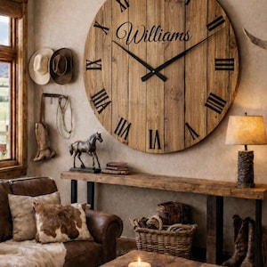 May include: Large, round wooden clock with black Roman numerals and hands. The clock face has the name "Williams" in script. The clock is mounted on a wall in a rustic living room setting. The text "clock with custom wording" is at the bottom.