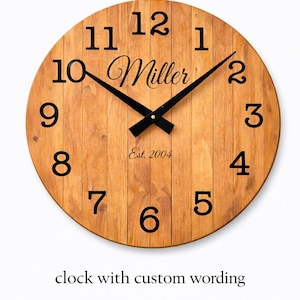 May include: A round wooden clock with black numerals and hands. The clock face has a light brown wood grain pattern. The word "Miller" is written in cursive, with "Est. 2004" below it. The text "clock with custom wording" is at the bottom.