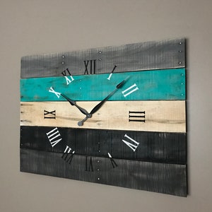 May include: A rustic wooden clock with Roman numeral markings on a multi-colored background. The clock face is painted in shades of gray, teal, and beige. The clock hands are black.