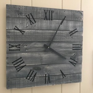 K&ouml;nnte beinhalten: Eine graue Holzuhr mit schwarzen r&ouml;mischen Ziffern und schwarzen Zeigern. Die Uhr ist an einer Wand montiert.