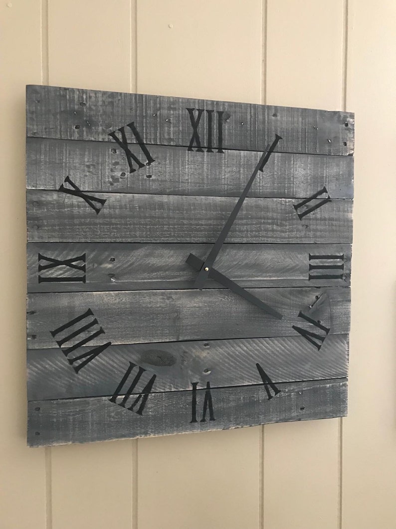 K&ouml;nnte beinhalten: Eine graue Holzuhr mit schwarzen r&ouml;mischen Ziffern und schwarzen Zeigern. Die Uhr ist an einer Wand montiert.
