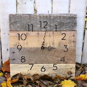 May include: A rustic wooden clock with black painted Roman numerals and hands. The clock is made from reclaimed wood planks and has a natural wood finish.