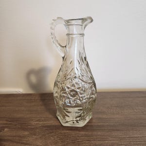 May include: A clear glass pitcher with a handle and a spout. The pitcher has a decorative, textured pattern on the body. The pitcher is sitting on a dark brown wooden surface.