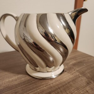 May include: Creamer with a white and silver swirl design. The creamer has a handle and a spout. The base and the rim of the spout are silver. The creamer is sitting on a wooden surface.