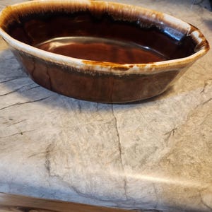 Puede incluir: Fuente de horno ovalada de cerámica marrón y blanca. La fuente tiene un acabado brillante con un interior marrón oscuro y un borde marrón claro y blanco. La fuente es probablemente vintage y adecuada para hornear y servir.