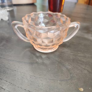 Puede incluir: Un azucarero vintage de cristal rosa claro con dos asas. El cuenco presenta un patrón geométrico en forma de diamante y un borde festoneado. El azucarero está sobre una superficie gris oscuro.