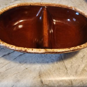 May include: A brown, oval-shaped divided serving dish with a glossy finish. The dish has a lighter, textured rim. The interior is separated by a vertical divider, creating two distinct compartments. This vintage piece is suitable for serving snacks or appetizers.