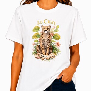 May include: A white t-shirt featuring a graphic print of a cheetah sitting among green leaves and pink flowers. Above the cheetah, the text reads LE CHAT in gold letters, and below the cheetah, a banner displays the word SAVAGE in black text.