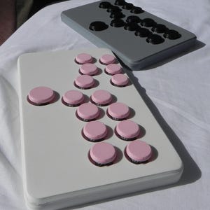 Könnte beinhalten: Zwei rechteckige Spielbretter, eines grau und eines weiß, mit kreisförmigen Knöpfen. Das graue Brett hat schwarze Knöpfe, die in einem Muster angeordnet sind. Das weiße Brett zeigt rosa Knöpfe in einem ähnlichen Layout. Die Bretter liegen auf einer weißen Oberfläche.