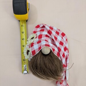 May include: A decorative gnome with a red and white checkered hat, brown beard, and wooden nose. The gnome is adorned with sunflower accents and measures approximately 7 inches in height, as indicated by the yellow measuring tape.