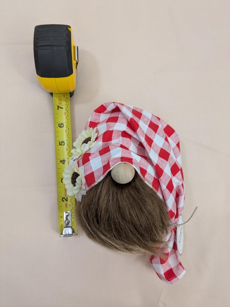 May include: A decorative gnome with a red and white checkered hat, brown beard, and wooden nose. The gnome is adorned with sunflower accents and measures approximately 7 inches in height, as indicated by the yellow measuring tape.