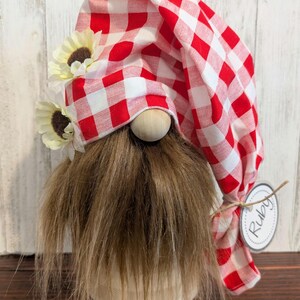 May include: A decorative gnome with a red and white checkered hat and a long, brown faux fur beard. The gnome has a wooden nose, cream-colored sunflowers, and a tag that says "Ruby". The gnome is on a cream-colored base.