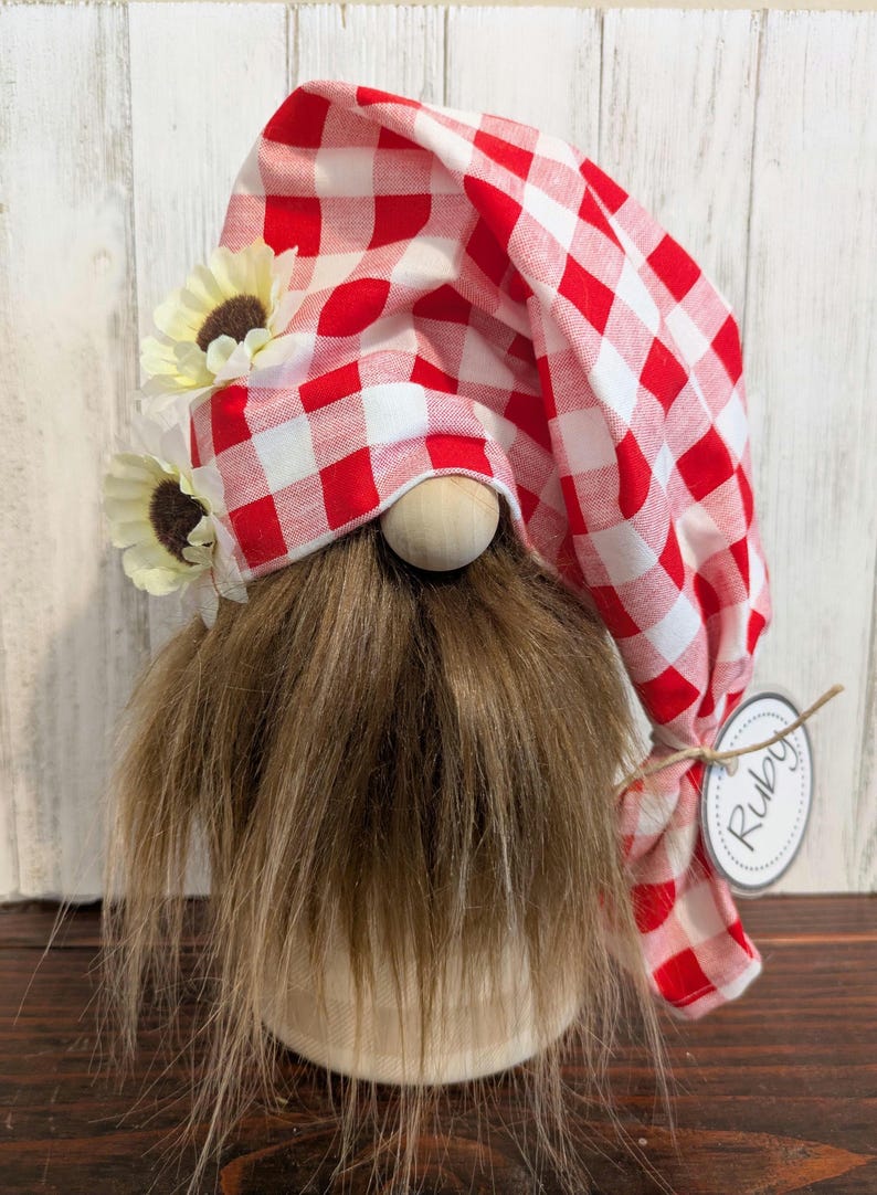May include: A decorative gnome with a red and white checkered hat and a long, brown faux fur beard. The gnome has a wooden nose, cream-colored sunflowers, and a tag that says "Ruby". The gnome is on a cream-colored base.