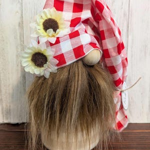 May include: A decorative gnome with a long brown beard and a red and white checkered hat. The hat is adorned with two small sunflowers. The gnome has a wooden nose and a beige body. A charming addition to any home decor.
