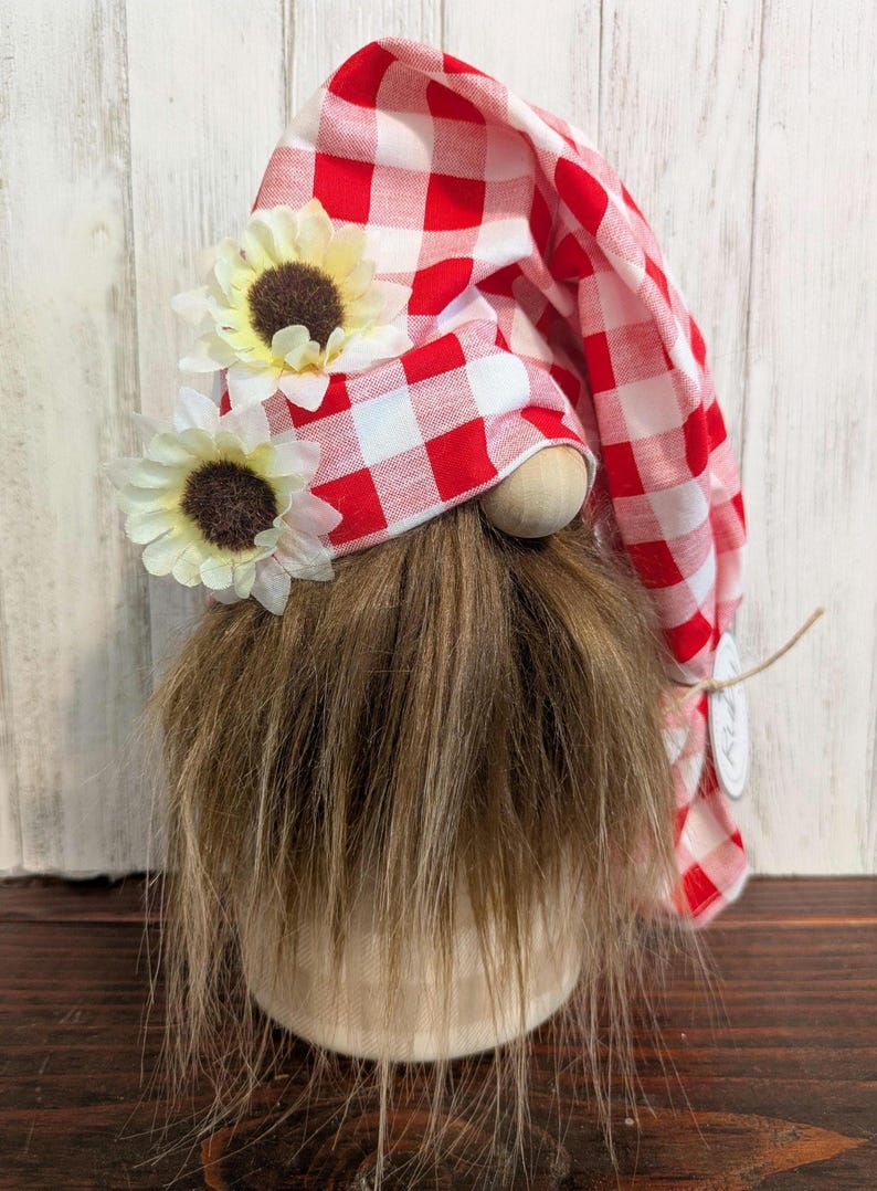 May include: A decorative gnome with a long brown beard and a red and white checkered hat. The hat is adorned with two small sunflowers. The gnome has a wooden nose and a beige body. A charming addition to any home decor.