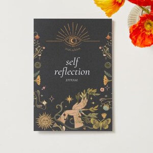 Peut inclure: Un journal noir avec les mots "self reflection" et "2026 edition" imprim&eacute;s sur la couverture. La couverture pr&eacute;sente des illustrations dor&eacute;es d'un &oelig;il, d'un soleil, d'&eacute;toiles et de papillons. Un vase de fleurs orange et jaunes est &agrave; droite.