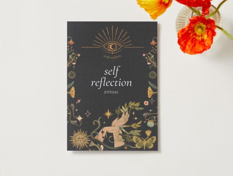 Peut inclure: Un journal noir avec les mots "self reflection" et "2026 edition" imprim&eacute;s sur la couverture. La couverture pr&eacute;sente des illustrations dor&eacute;es d'un &oelig;il, d'un soleil, d'&eacute;toiles et de papillons. Un vase de fleurs orange et jaunes est &agrave; droite.