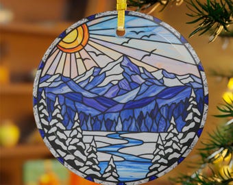 May include: Round ceramic ornament featuring a stained-glass design of a mountain landscape. The design includes a sun, mountains, trees, and a river, with a color palette of blues, white, and orange. A gold ribbon is attached for hanging.