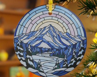 May include: Round Christmas ornament with a mountain landscape design. The ornament features a stained-glass style image of snow-covered mountains, a river, and evergreen trees, with a rainbow and sun in the background. It hangs from a gold ribbon.