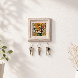 Könnte beinhalten: Ein quadratischer Wand-Schlüsselhalter mit einem weiß gewischten Holzrahmen, der einen Blumendruck mit gelben und weißen Blüten in einer grünen Vase umgibt. Drei goldfarbene Metallhaken sind unten befestigt und halten jeweils einen Metallschlüsselbund.