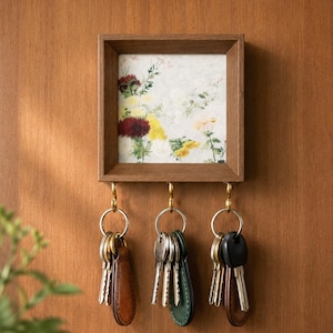 Puede incluir: Un soporte para llaves de pared de madera cuadrada con un inserto de estampado floral que muestra ilustraciones botánicas en rojo, amarillo y verde. Tres ganchos de metal color latón sobresalen de la parte inferior, cada uno con un llavero plateado.