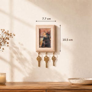 Könnte beinhalten: Ein rechteckiger Schlüsselhalter aus hellem Holz zur Wandmontage mit einem kleinen Bilderrahmen und drei Messinghaken für Schlüssel. Der Rahmen ist 7,7 cm breit und 10,5 cm hoch und bietet eine funktionale Aufbewahrungsmöglichkeit für Schlüssel.