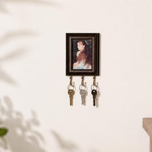 Könnte beinhalten: Ein rechteckiger, dunkelbrauner Schlüsselhalter aus Holz zur Wandmontage mit einem kleinen Porträt einer Person mit langem Haar in einem Rahmen. An der Unterkante befinden sich drei Metallhaken, an denen jeweils ein Metallschlüssel hängt.