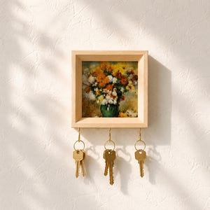 Könnte beinhalten: Ein quadratischer Schlüsselhalter aus hellem Holz mit einem gerahmten Blumenbild, das gelbe, orange und weiße Blüten in einer grünen Vase zeigt. An der Unterkante befinden sich drei goldene Metallhaken, an denen jeweils Schlüssel hängen.