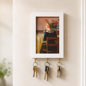 Könnte beinhalten: Ein weiß gerahmter Schlüsselhalter mit einem Vintage-Gemälde einer Person, die an einem Tisch mit Blumen sitzt. Der Schlüsselhalter hat drei Messinghaken mit daran hängenden Schlüsseln. Er ist an einer cremefarbenen Wand befestigt.