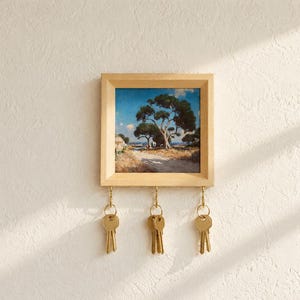 Könnte beinhalten: Ein quadratischer Schlüsselhalter aus Holz mit einem gerahmten Landschaftsgemälde von Bäumen unter blauem Himmel. Der helle Holzrahmen hat drei goldfarbene Metallhaken an der Unterkante, an denen jeweils ein Schlüsselbund aus Silber hängt.