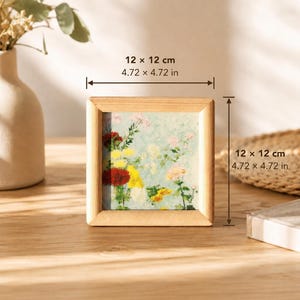 Puede incluir: Un marco de madera cuadrado que contiene un estampado floral con flores rojas, amarillas y rosas sobre un fondo azul claro. El marco mide 12 por 12 cm, como indican las líneas de medición y el texto impreso encima y al lado del objeto.