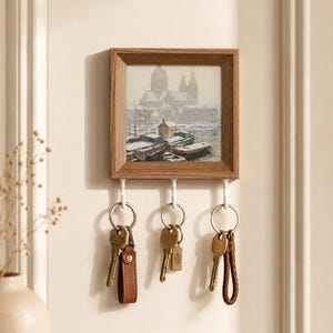 May include: A square wooden wall-mounted key holder featuring a framed print of a snowy harbour scene with boats. The unit includes three white hooks, each holding a metal keyring with various attached keys and leather or braided key fobs.