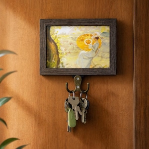 May include: A framed artwork depicting a woman with a yellow hat, hanging above a key holder with several keys. The frame is dark brown, and the key holder is bronze. The artwork is mounted on a wooden door.