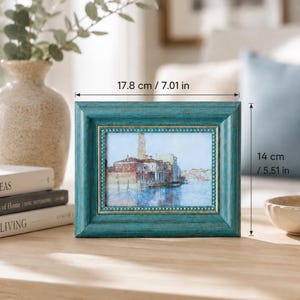 Può includere: Una cornice rettangolare color verde acqua con finitura anticata e bordo interno dorato, contenente un dipinto paesaggistico di una città sull'acqua. La cornice misura 17,8 cm di larghezza e 14 cm di altezza come indicato dalle etichette.