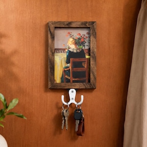 May include: A framed artwork depicting a person seated at a table with flowers, mounted on a wall with a white key hook. Keys hang from the hook. A coat and hat hang on the wall to the right. A plant in a vase is on the left.