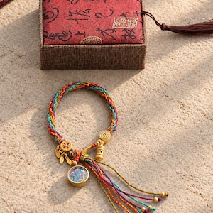 Può includere: Un bracciale in corda intrecciata multicolore con ciondoli in metallo dorato, tra cui un pendente circolare con inserti colorati, piccoli ciondoli a forma di foglia e una perla cilindrica, accompagnato da una scatola in tessuto rosso con calligrafia.