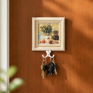 Può includere: Un dipinto con cornice di natura morta di fiori e frutta è appeso sopra un gancio per chiavi bianco. Il gancio contiene un set di chiavi e un portachiavi per auto. La cornice è di colore chiaro con un bordo decorativo, montata su una porta di legno marrone.