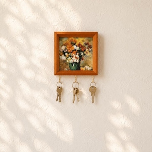 Könnte beinhalten: Ein quadratischer Schlüsselhalter aus Holz mit einem gerahmten Druck eines Blumenstraußes in einer grünen Vase. Der Rahmen hat drei goldfarbene Metallhaken an der Unterkante, an denen jeweils ein silberner Metallschlüssel an einem Ring hängt.