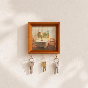 Könnte beinhalten: Ein quadratischer Schlüsselhalter aus Holz mit einem gerahmten Druck einer Innenszene mit Tisch, Stühlen und Lampe. An der Unterkante des Rahmens befinden sich drei messingfarbene Metallhaken, an denen jeweils ein Schlüsselbund hängt.