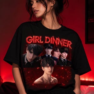 Può includere: T-shirt nero con la scritta "GIRL DINNER" in lettere glitterate rosse. La maglietta presenta una grafica di diversi personaggi maschili in stile anime in varie pose. Lo sfondo è rosso scuro con piccole forme di cuore.