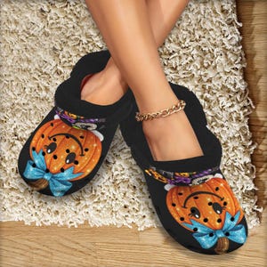 May include: A pair of black slip-on clogs featuring a graphic print of a smiling orange pumpkin with a glittery blue bow. The shoes have a black interior lining and a rounded toe design, showcasing a festive seasonal pattern on the outer surface.