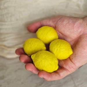 Könnte beinhalten: Vier kleine, leuchtend gelbe, zitronenförmige Objekte aus gefilzter Wolle. Jedes Stück hat eine texturierte, flauschige Oberfläche und eine ausgeprägte ovale Form mit einem kleinen spitzen Ende, die wie Miniaturzitronen aussieht.