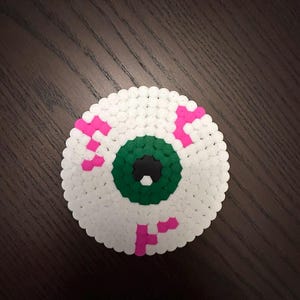 Op de afbeelding: Een cirkelvormig oogontwerp gemaakt van witte, groene, roze en zwarte kralen. Het oog heeft een groene iris, een zwarte pupil en roze aderen. Het ontwerp is geplaatst op een donkerbruin houten oppervlak.