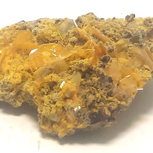 Puede incluir: Un espécimen mineral con un grupo de cristales translúcidos de color naranja sobre una matriz de roca marrón oscuro y amarilla. Los cristales tienen una apariencia vítrea, reflejando la luz. La textura general es áspera y desigual.