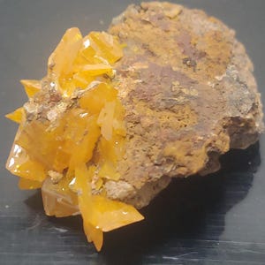 May include: A mineral specimen featuring a cluster of vibrant, translucent orange crystals growing from a rough, textured, brownish-gray rock matrix. The crystalline structure displays sharp, angular facets that catch the light, contrasting with the earthy, uneven surface of the host stone.